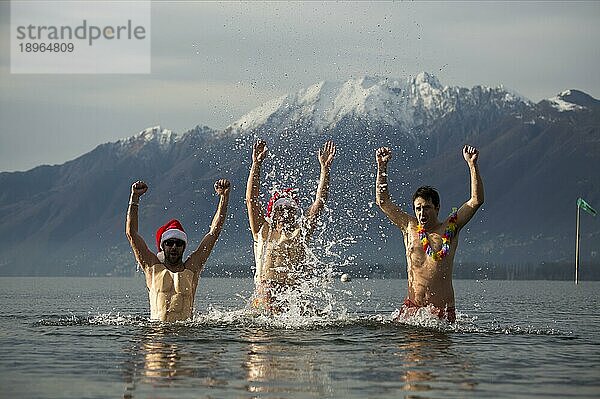 Drei glückliche Weihnachtsmänner mit ausgestreckten Armen in einem kalten alpinen Lago Maggiore mit schneebedecktem Berg in Locarno  Schweiz  Europa