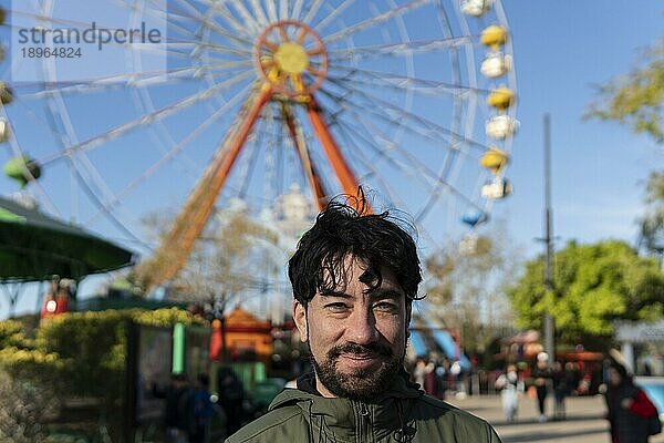 Porträt eines Latinomannes in einem Vergnügungspark  der glücklich mit dem Riesenrad im Hintergrund posiert