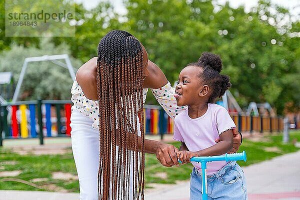 Schwarzafrikanische Mutter hat Spaß mit ihrer Tochter auf dem Spielplatz  die mit dem Skateboard fährt und ihre Zunge herausstreckt