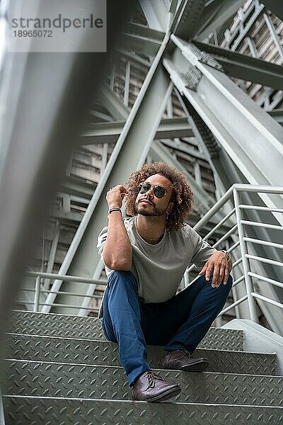 Junger Mann mit Afro Haare tragen Sonnenbrille auf der Treppe in der Stadt sitzen  urbane Mode cooles Konzept eines Hipster Kerl mit stilvollen