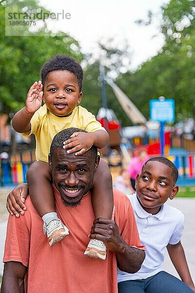 Porträt eines afrikanischen Vaters schwarzer Herkunft  der mit seinen Kindern auf einem Spielplatz im Stadtpark lächelt