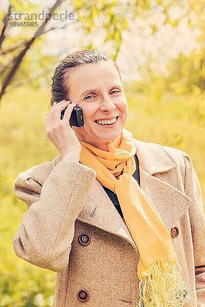 Porträt einer reifen und lächelnden Geschäftsfrau  die einen Mantel und einen gelben Schal trägt und mit ihrem Mobiltelefon telefoniert