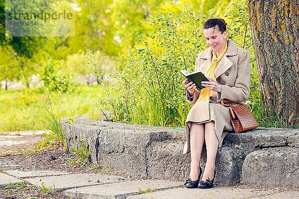 Elegante Geschäftsfrau  die an einem sonnigen Frühlingstag auf einer Steinmauer sitzt und ein lustiges Buch liest