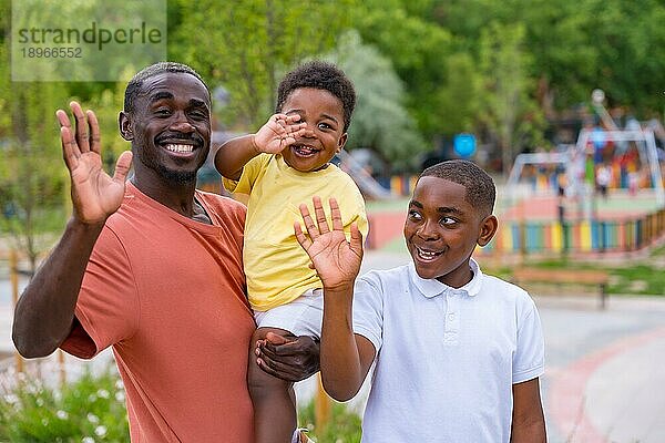 Schwarzafrikanischer Vater hat Spaß mit seinem Sohn auf dem Spielplatz und winkt in die Kamera