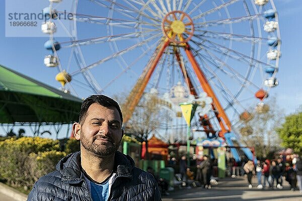 Porträt eines Latinomannes in einem Vergnügungspark  der glücklich mit dem Riesenrad im Hintergrund posiert