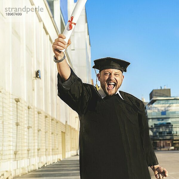 Glücklicher erwachsener Mann mit Bachelor Kleid und schwarzem Mörtelbrett  der sein Diplom zeigt und in die Kamera schaut