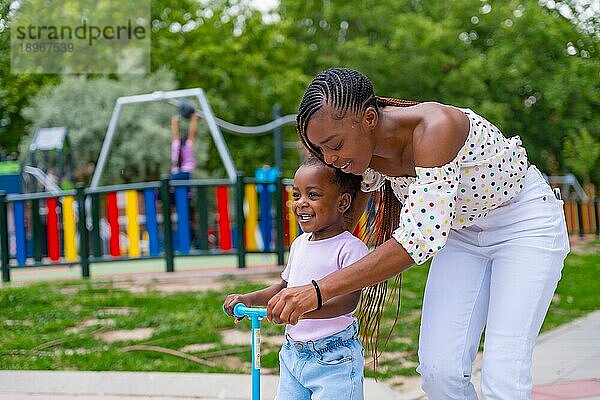 Schwarzafrikanische Mutter hat Spaß mit ihrer Tochter auf dem Spielplatz und lernt  Skateboard zu fahren