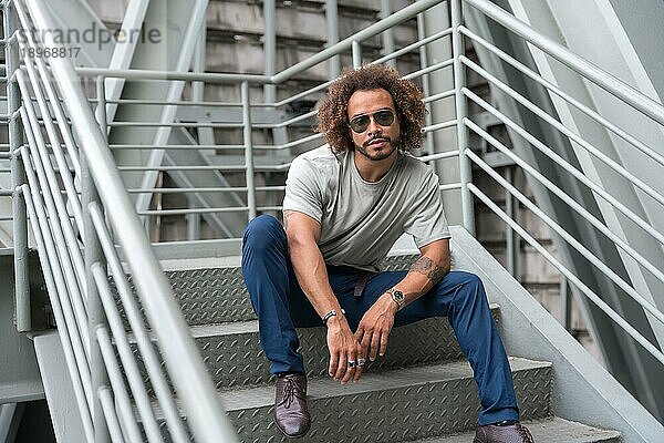 Junger Mann mit Afro Haare tragen Sonnenbrille auf der Treppe in der Stadt sitzen  urbane Mode cooles Konzept eines Hipster Kerl mit stilvollen