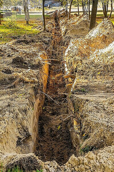 Ein Graben in der Erde für die Verlegung eines Rohrs im Park  unter den Bäumen  im abendlichen Herbstlicht