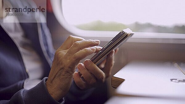 Nahaufnahme der Hände einer älteren Frau  die in einem Zugwaggon sitzt und ein Smartphone benutzt