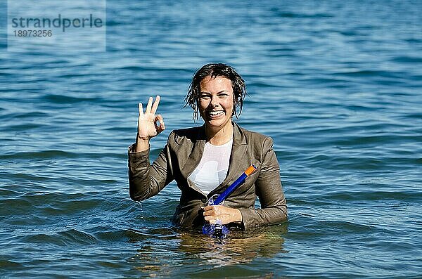 Elegante und glückliche nasse Frau im Wasser mit Tauchermaske und Anzug und zeigt das Ok Signal