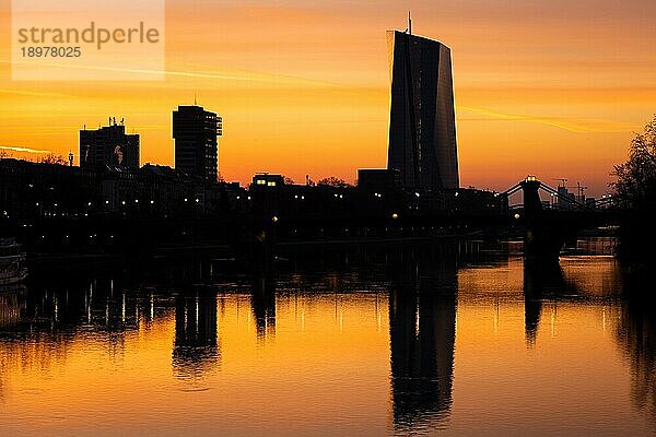 Die Silhouette der Europäischen Zentralbank (EZB) in Frankfurt am Main zeichnet sich kurz vor Sonnenaufgang gegen den gelb-orangenen Morgenhimmel ab und spiegelt sich im Main.  Frankfurt am Main  Hessen  Deutschland  Europa