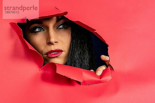 Wunderschönes brünettes Modell reißt durch rosa Papier und zeigt nur ihr Gesicht  Nägel und Lippen