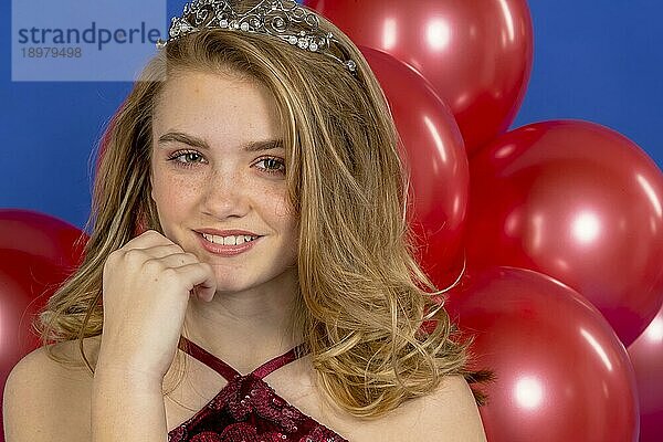 Ein schönes blondes Teenager Model posiert mit einem Diadem und roten Luftballons vor der Kamera in einer Studioumgebung