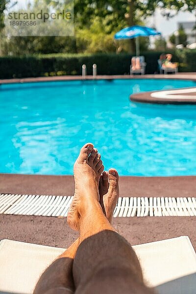 Die Beine eines Mannes  der sich auf einem Liegestuhl mit Blick auf das Wasser entspannt und ein Sonnenbad am Pool nimmt