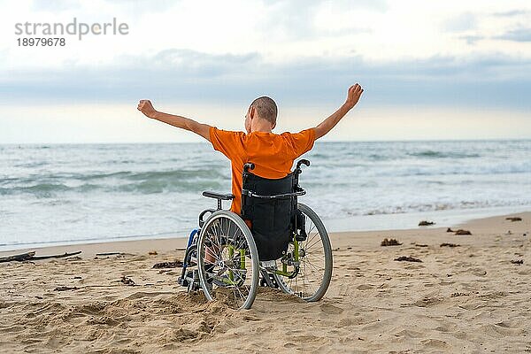 Behinderte Person auf dem Rücken in einem Rollstuhl am Strand mit offenen Armen  die die Freiheit genießen