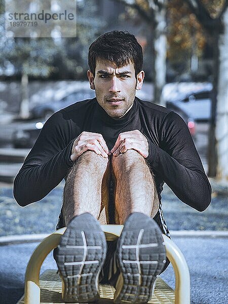 Vorderansicht eines jungen Mannes bei Bauchübungen an einem öffentlichen Fitnessgerät im Freien