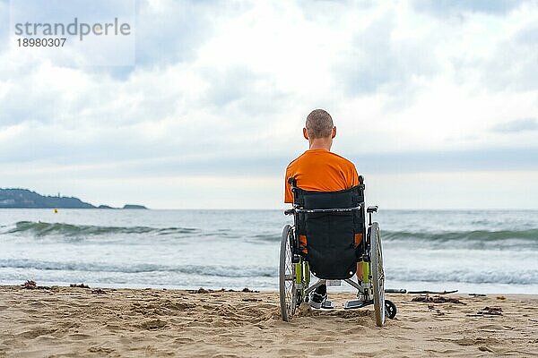 Ein Behinderter in einem Rollstuhl auf dem Rücken am Strand im Sommer mit Blick auf das Meer