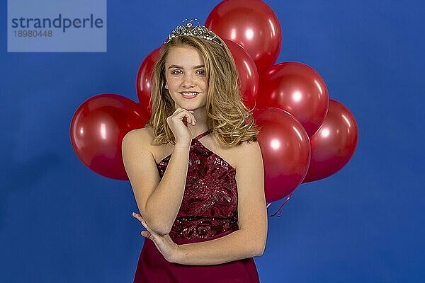 Ein schönes blondes Teenager Model posiert mit einem Diadem und roten Luftballons vor der Kamera in einer Studioumgebung