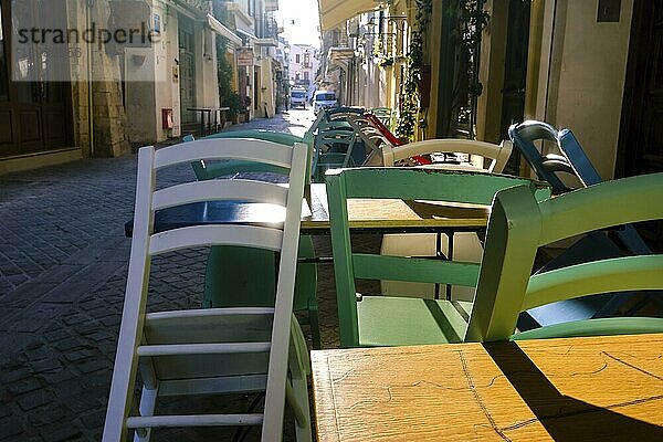 Früher Morgen in den leeren engen Straßen einer alten Mittelmeerstadt. Helles Sonnenlicht. Bunte Stühle und Tische in Cafés und Restaurants nicht bereit. Selektiver Fokus auf die Straßenmöbel im Vordergrund. Chania  Kreta  Griechenland  Europa