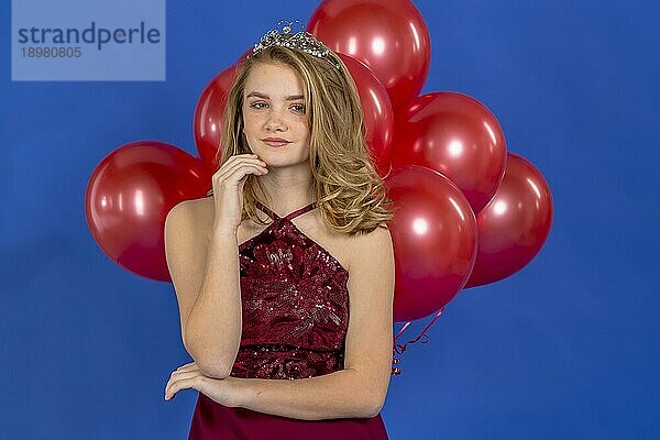 Ein schönes blondes Teenager Model posiert mit einem Diadem und roten Luftballons vor der Kamera in einer Studioumgebung