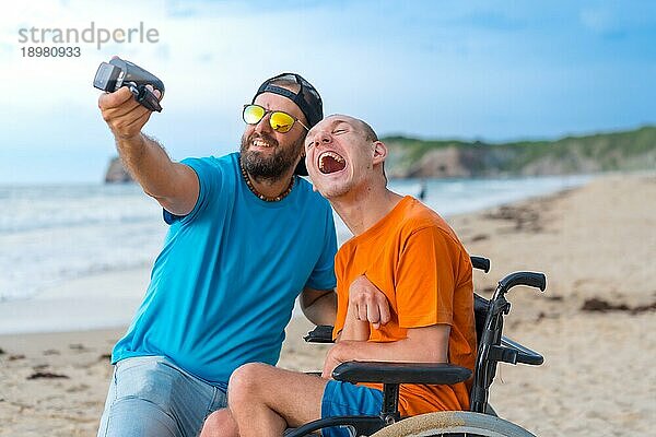 Eine behinderte Person im Rollstuhl am Strand mit einem Freund  der Spaß daran hat  ein Selfie zu machen