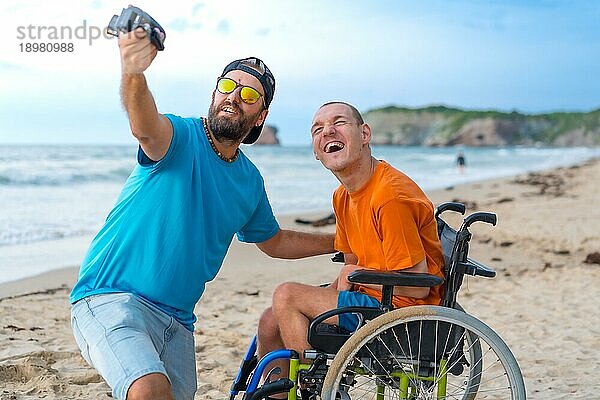 Eine behinderte Person in einem Rollstuhl am Strand mit einem Freund  der ein Selfie macht