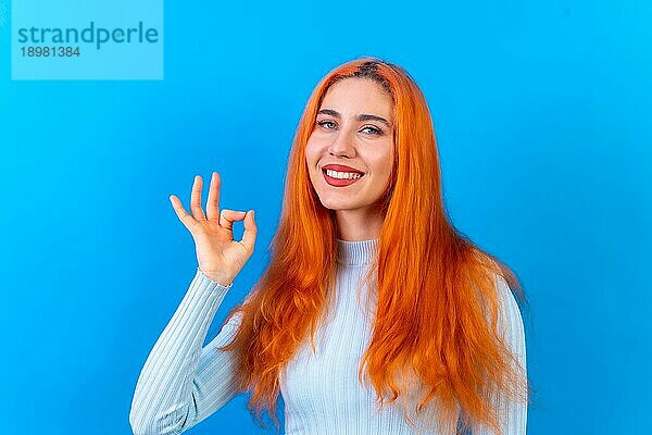 Rothaarige Frau im Studio Fotografie macht die ok Geste auf einem blauen Hintergrund