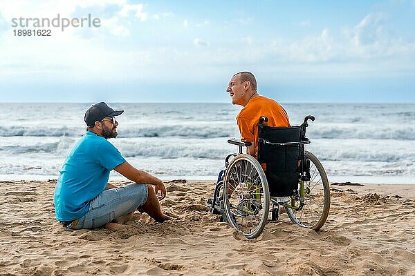 Porträt einer behinderten Person in einem Rollstuhl am Strand mit einem Freund  der auf das Meer blickt