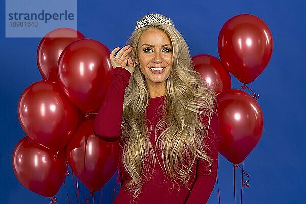 Ein schönes blondes Modell posiert mit einem Diadem und roten Luftballons vor der Kamera in einer Studioumgebung