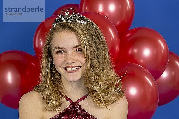 Ein schönes blondes Teenager Model posiert mit einem Diadem und roten Luftballons vor der Kamera in einer Studioumgebung