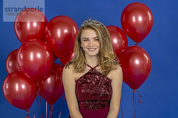 Ein schönes blondes Teenager Model posiert mit einem Diadem und roten Luftballons vor der Kamera in einer Studioumgebung