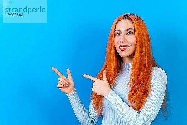 Rothaarige Frau in Studiofotografie macht die Geste des Zeigens auf einem blauen Hintergrund