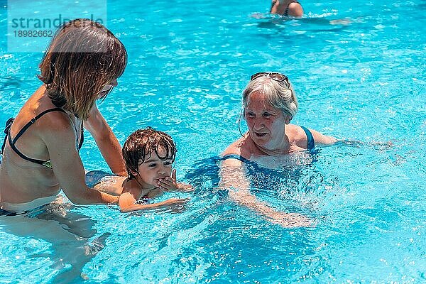 Mutter und Großmutter amüsieren sich im Sommerurlaub mit ihrem Enkel im Pool und lernen schwimmen