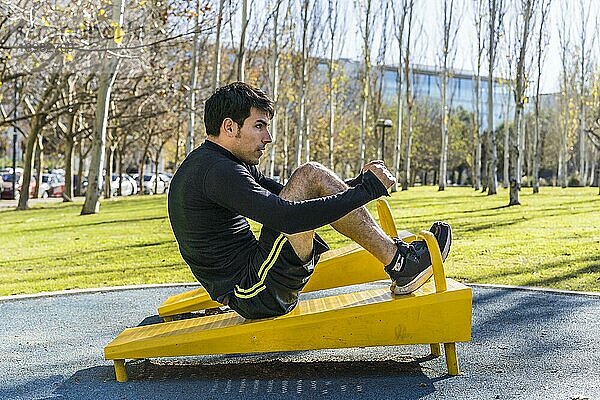 Selbstbewusster junger Mann bei Bauchmuskelübungen an einem öffentlichen Fitnessgerät im Freien
