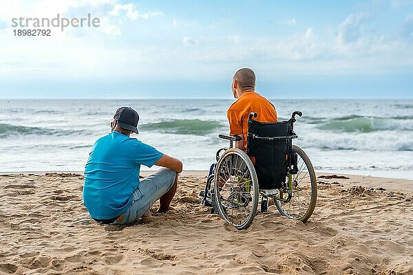 Porträt einer behinderten Person in einem Rollstuhl am Strand mit einem Freund  der auf das Meer blickt