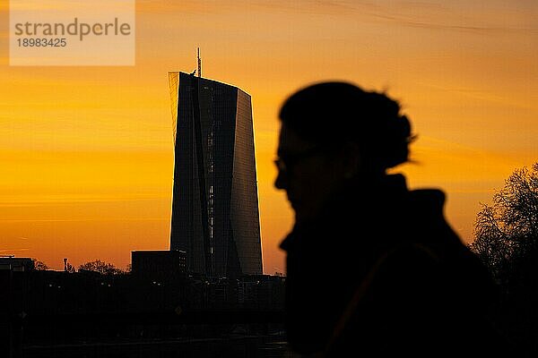 Die Silhouetten der Europäischen Zentralbank (EZB) in Frankfurt am Main und einer Passantin zeichnen sich kurz vor Sonnenaufgang gegen den gelb-orangenen Morgenhimmel ab.  Frankfurt am Main  Hessen  Deutschland  Europa