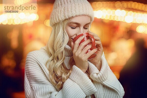 Junge Frau  die eine Tasse heißen Kaffee im Winter genießt und ihn in ihren Händen schröpft  während sie mit einem seligen Lächeln vor einem glühend warmen orangefarbenen Hintergrund trinkt