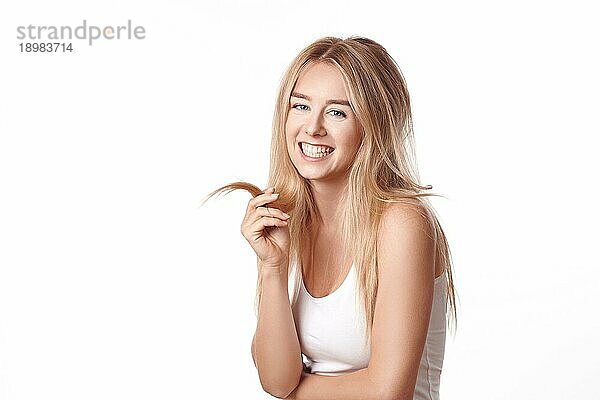 Attraktive junge blonde Frau mit einem freundlichen glücklichen Lächeln  die in die Kamera grinst  während sie mit einer Strähne ihres langen Haares spielt  vor weißem Hintergrund