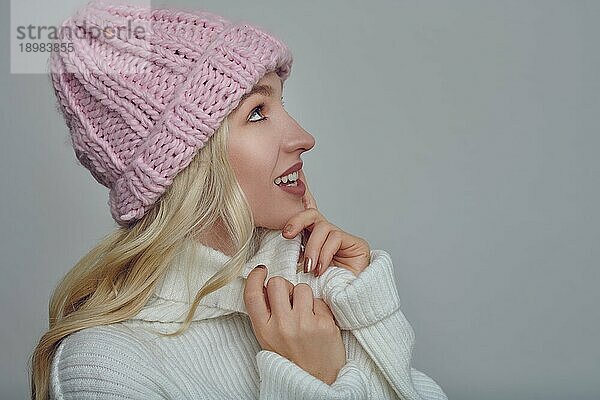 Hübsche junge blonde Frau in Woll Winterkleidung  die sich mit einem glücklichen Lächeln in die Wärme ihres Rollkragenpullovers kuschelt  während sie eine rosa Strickmütze trägt  Profilporträt in Grau mit Leerzeichen