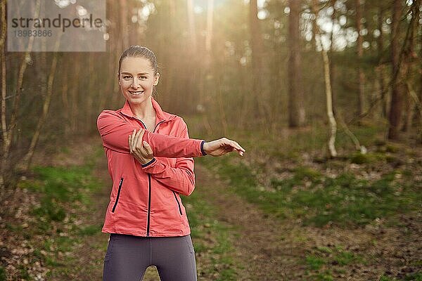 Frontansicht einer glücklichen  fitten Frau  die ihren Arm bei Aufwärmübungen im Freien im Wald im Frühling ausstreckt