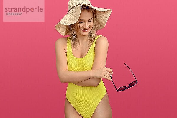 Schlanke attraktive junge blonde Frau in einem bunten gelben Badeanzug und mit einem breitkrempigen Strohsonnenhut  der mit einem glücklichen Lächeln zur Seite schaut  während sie ihre Sonnenbrille auf rosa Hintergrund hält