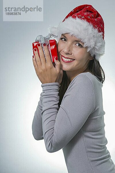 Schöne Frau mit roter Nikolausmütze  die mit einem charmanten  freundlichen Lächeln ein verpacktes Geschenk an die Wange hält  auf grauem Hintergrund mit Platz für Ihren Weihnachtsgruß