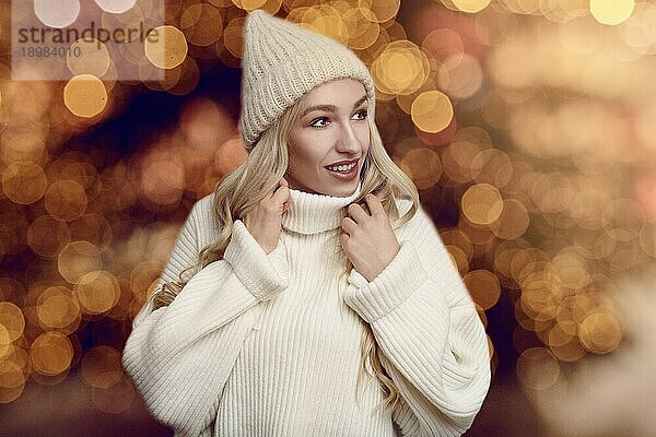 Schöne junge blonde Frau in weißem Strickpulli und Wintermütze  schaut in die Kamera und lächelt  steht vor warmen gelben Lichtern mit Bokeheffekt verschwommen