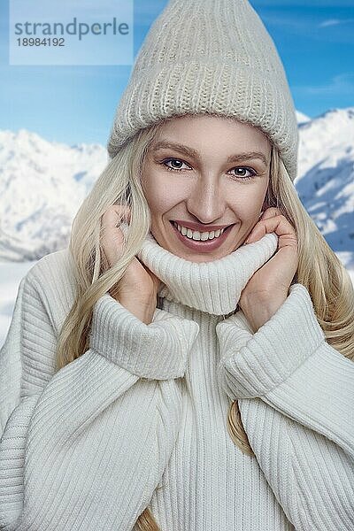 Attraktive blonde Frau in einem warmen Wollpullover mit Rollkragen und Strickmütze steht im Freien und kuschelt sich mit einem breiten  glücklichen und freundlichen Lächeln in den Kragen vor verschneiten Berggipfeln