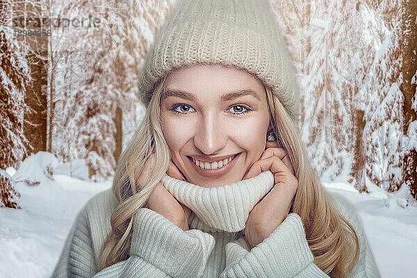 Attraktive blonde Frau in einem warmen Wollpullover mit Rollkragen und Strickmütze steht im Freien und kuschelt sich mit einem breiten  glücklichen und freundlichen Lächeln an einen verschneiten Waldweg