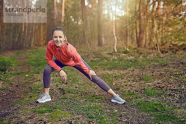 Ganzkörperporträt einer glücklichen  fitten jungen Frau  die Dehnübungen für Beine und Arme während eines Trainings im Freien im Wald im Frühling macht