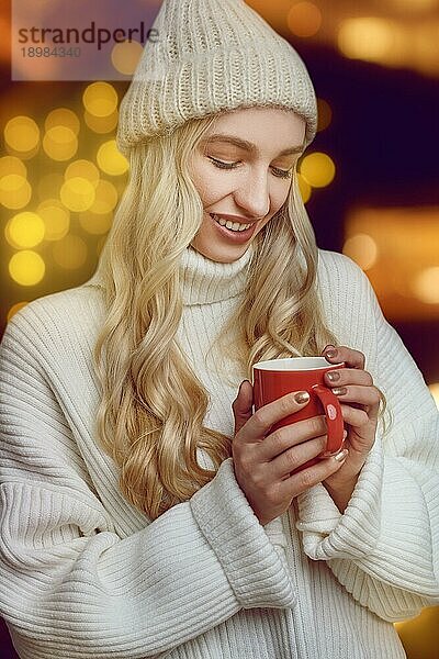 Junge Frau  die eine Tasse heißen Kaffee im Winter genießt und ihn in ihren Händen schröpft  während sie mit einem seligen Lächeln vor einem glühend warmen orangefarbenen Hintergrund trinkt
