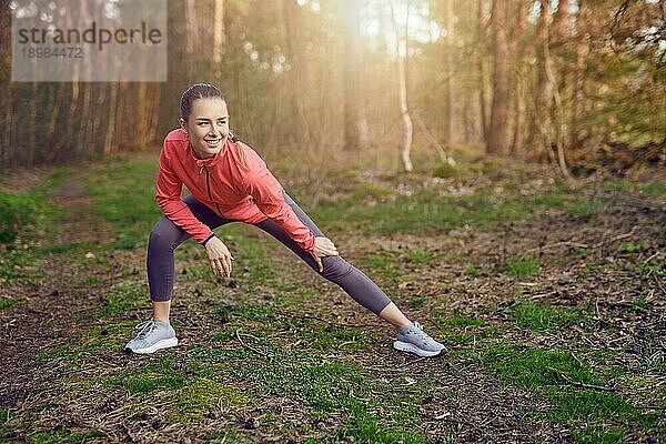 Ganzkörperporträt einer glücklichen  fitten jungen Frau  die Dehnübungen für Beine und Arme während eines Trainings im Freien im Wald im Frühling macht