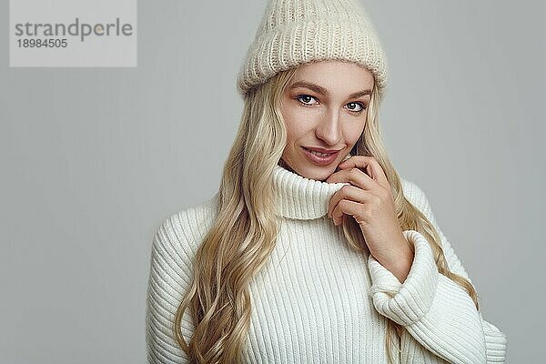 Schöne junge blonde Frau in weißem Strickpulli und Hut lächelt und schaut in die Kamera. Nahaufnahme Frontalporträt gegen einfachen hellblauen Hintergrund